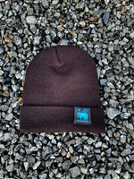 Marin knit beanie with embroidered tag, black, on gravel.